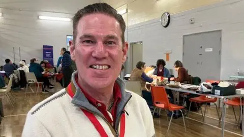 Stuart King is centre frame and smiling to the camera. He has short dark hair and is wearing a white, half zip jumper over a red polo shirt. He has a red lanyard around his neck. He is in a community centre hall, and behind him a repair cafe is in action. There are groups of people at tables focusing on different repairs and working.