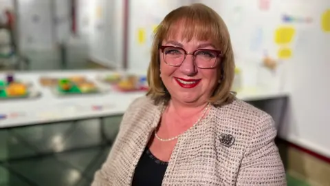 Sharon Hodgson smiling into the camera, she has light, short hair with a full fringe. She is wearing large glasses, red lipstick and a pearl necklace along with a silver patterned blazer.