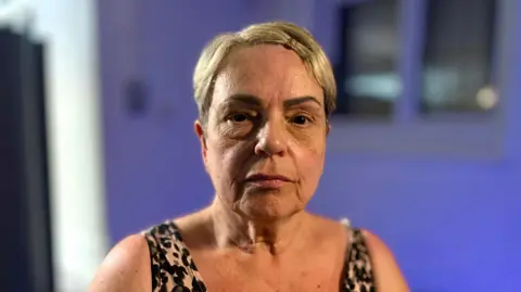 MARTIN GILES/BBC A head and shoulders picture of Linda Stokes who is looking at the camera with a straight face. Behind her the background is blurry. It appears to be a white wall with blue light shining on it and a window. 
