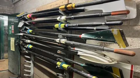 Several oars are stacked on small metal shelving on the wall of a clubhouse. They are covered in dirt and mud from flood water