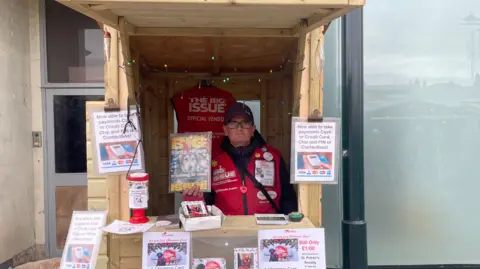 Big Issue seller in wooden shelter 