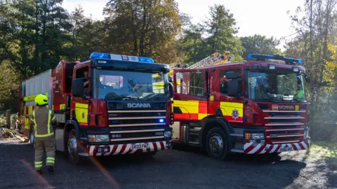 Two fire service vehicles parked on tarmac. Both are red with yellow high-vis panels and blue emergency lights above the cab which are not switched on. One has high reaching ladders on its back. A firefighter is seen approaching the other. There are trees in the background and the sun is shining