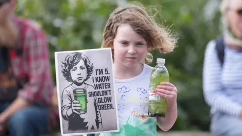 PA Media A five-year-old girl with long brown hair and a white t-shirt holding a bottle of green water from the lough and a poster.