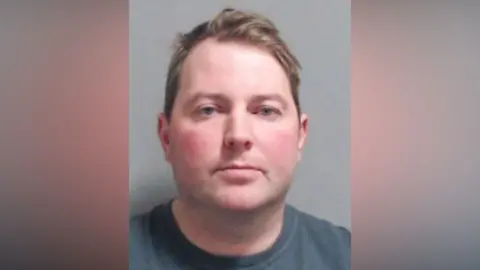 PSNI Jonathan Creswell has short brown hair parted to the side and light blue eyes. He is wearing a blue/grey t-shirt and is staring straight ahead in a police mugshot.