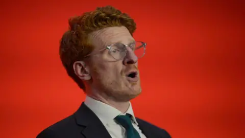 PA Matthew is mid speech in front of a bold red background. 
He is wearing silver small rimmed glasses with a pink arm. 
He has ginger curly short hair.
He's wearing a teal blue tie, white shirt and navy suit blazer. 
