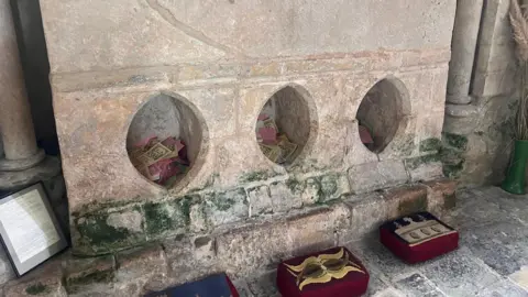Three large oval holes are carved into the marble beneath a shrine. Before them are three kneelers on the floor.