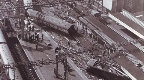 Walter Bloxham/Warwickshire Police/Nuneaton Memories A black and white aerial view of the aftermath of the crash with railway tracks visible between two stations. Train carriages can be seen, strewn across the tracks with two of them having come uncoupled.