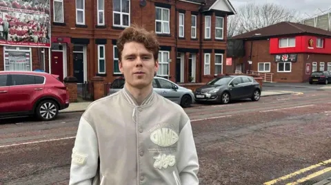 Ginger-haired man in grey/cream baseball jacket stands on roadside near Old Trafford