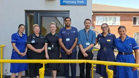 Royal Wolverhampton NHS Trust A group of seven medical professionals stand in a line in their scrubs, smiling at the camera