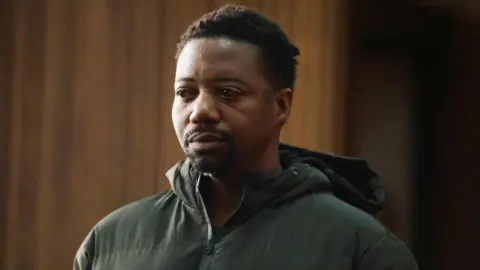 A man in dock in court. He is wearing a dark green coat with a hood. 