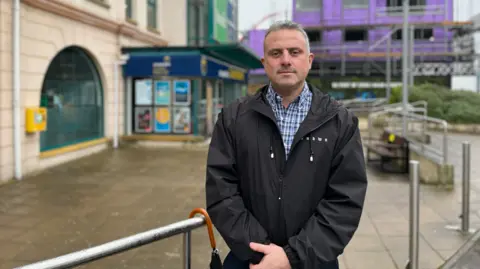 Carl Walker, a man with grey hair and grey stubble. He is wearing a shirt with blue and white checks, underneath a black hooded coat with a zip through the middle. He is standing outside a Coop supermarket in St Helier.