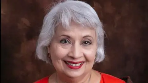 A caput and shoulders image of a female pinch grey hairsbreadth smiling astatine nan camera successful a workplace portrait. Her hairsbreadth is successful a bob style pinch a fringe. She is wearing reddish lipstick and a reddish top. The inheritance is simply a mottled brown.