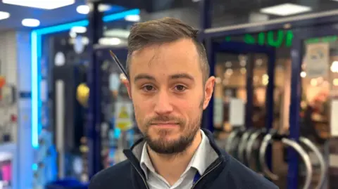 Man with brown hair and beard, wearing a blue shirt and zipped jumper is smiling at camera. He has a pen sat behind his ear. The front doors to his store can be seen behind him. 