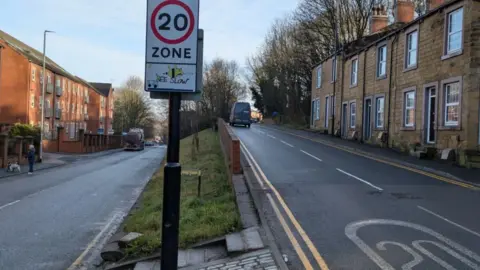 A fork in a road with a 20mph roadsign between the two parallel roads. One of the roads leads uphill with terraced houses on the right.