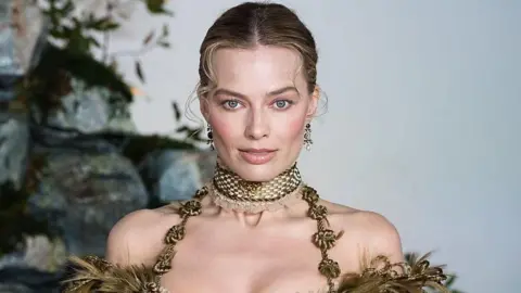 Getty Images Margot Robbie has her blonde hair swept back with two curly tendrils at the front. She has dangly earrings and a gold choker-style thick necklace