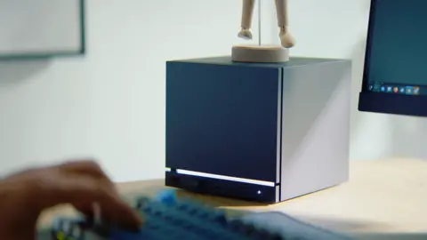 Valve Steam Machine sits on a wooden desk. Someone's hand is typing on a keyboard, in front of a PC monitor, beside it.