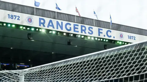 SNS General view of Ibrox stadium under a grey sky showing the top of the goal and the main stand with flags fluttering in the wind above it
