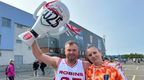 Chris wears a Robins white and red vest and holds up a giant blow up trophy with the Robins 2025 on it.  His arm is around his female friend.