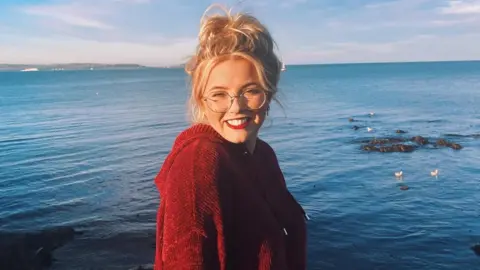Alexandra Galbraith A woman with blonde hair and glasses smiling at the camera in front of the sea, she is wearing a red hoodie