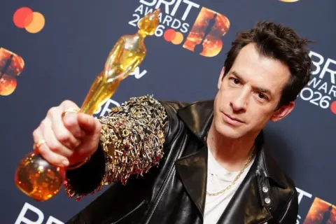 PA Media Mark Ronson holds his award up to the camera. He is wearing a leather jacket from the Celine collection.