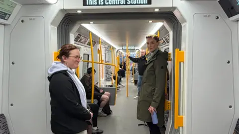 Two undercover police officers stand in between two Metro train carriages. One of them (left) is wearing a grey hoody, and grey sweatpants, and has a small black earpiece in her ear. She is smiling at a passenger who is sitting down opposite her. On the right, her colleague is wearing a long green jacket and holding a purple metal water bottle. The Metro train is in motion and inside a black tunnel.
