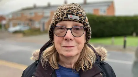 Sam Read/BBC Ann Nevison standing in front of a row of houses and a green bus, which is slightly blurred. She is wearing a leopard print hat, with glasses and shoulder-length fair hair. She is wearing a black coat, with fur trim, a blue top and jumper.