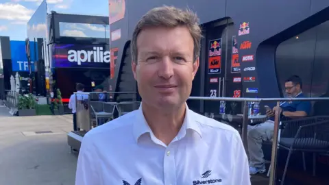 James Burridge/BBC Stuart Pringle is wearing a white Silverstone branded shirt and smiles at the camera. He is standing next to branded buildings at the track.