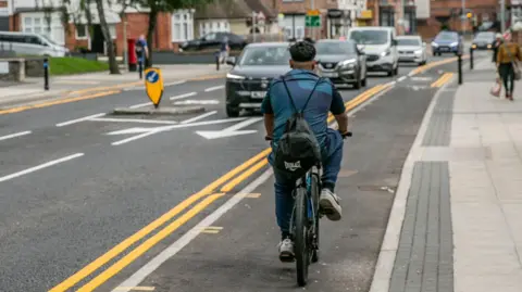 Rear view of a man cycling. He is riding along a dedicated cycle path with cars approaching him in the opposite direction. The setting is a residential street, with houses on the visible left side, and pedestrians on both pavements.