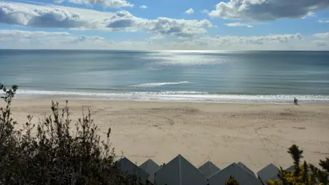 LouieMurg A wide view of a beach with just two people walking along the sand in the distance and the blue sea and sky in the background. 