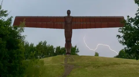 Simon The imposing, rust-coloured Angel of the North statue stands on top of a small, grassy hill. The sky is grey and stormy. A fork of lightning can be seen under the angel's right wing. 
