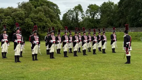 20 men in full ceremonial dress, including military style jackets and hats with feather plumes. The uniforms are navy, with white and red stripes across the front. They are wearing black boots and have swords on their sides.