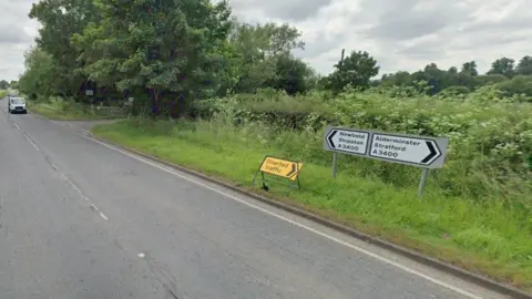 A rural road, with signs pointing on the A3400 towards Newbold and Shipston to the left, and A3400 Alderminster Stratford to the right. Trees line the road. A grey car can be seen travelling from left to right.