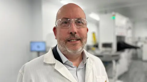 Tony Pearce looking into camera with a technology lab behind him. He is bald, with a grey closely-shaved beard and is wearing a lab coat over a suit and shirt.