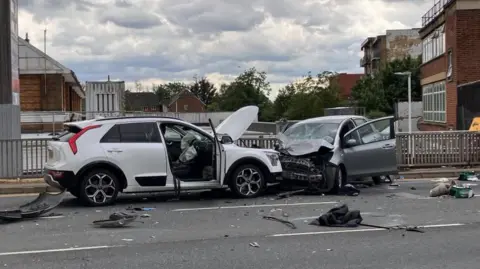 A white car and a grey car are badly damaged from the impact with debris scattered across the road.