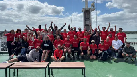 Saints Foundation A group of Benali on Tour participants on the ferry some stood and some sat cheering at the camera. All are wearing red t-shirts and Franny Benali is visible in the centre