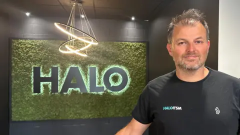 Alice Cunningham/BBC Paul Hamilton stands and smiles at the camera in front of a large sign that reads his company's name, Halo. He has grey/dark hair and wears a black T-shirt with his company's ITSM logo on it.