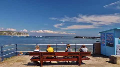 Robin Red Breast Four people sat on a bench looking out at the sea with clouds in the blue sky and a boats on the water. The pier is visible in the distance.