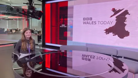 Mia is sat in the presenting chair of the Wales Today studio. she is smiling at the camera sat behind the desk. Behind her is the screen which shows BBC Wales Today