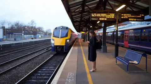 Getty Images A woman in a dark coat and high heels is standing on a train station platform looking at her phone as a train is coming in.  Above her are electronic boards displaying train times.