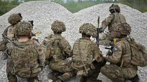 Six soldiers wearing camouflage face away from the camera, kneeling in pile of grey stones while on exercise