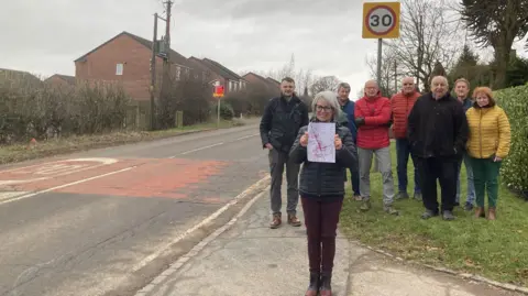 Eight people gathered at the side of the road at the entrance of a village, underneath a 30mph sign. A woman standing in front holds up a piece of paper with a diagram on it.