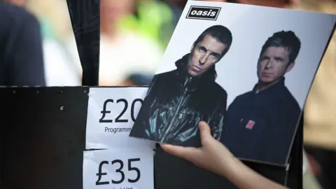 A hand holds up a photo of Liam and Noel Gallagher on a record in front of a sign which says £20 programmes and £35