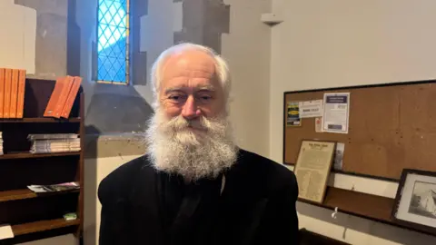 BBC A man with white hair and long bushy white facial hair is wearing a black jacket and is standing inside a church