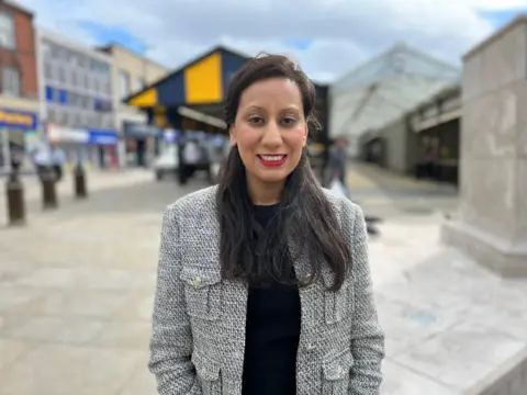 A woman wearing a grey blazer and a black top. She has dark hair and red lipstick.