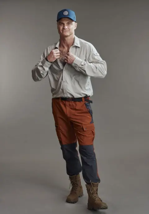 Channel 4 Promotion image of Tomasz for the Channel 4 show Tempting Fortune. He is wearing a baseball cap, a beige shirt, orange and black cargo trousers and brown hiking boots.