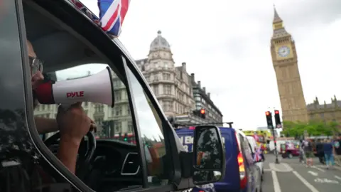 BBC A van driver speaks through a megaphone in phone of Big Ben as other vans queu in the distance