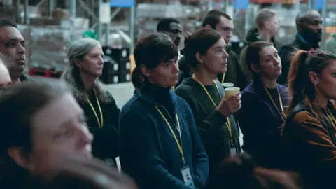 Conic Film A woman stands in the centre of a group of warehouse workers. She is wearing a dark blue jumper and is wearing a lanyard with her ID on it, similar to the others. They are staring all in the same direction, presumably paying attention to a manager giving a speech, but the expression on her face shows she might be in her own thoughts.