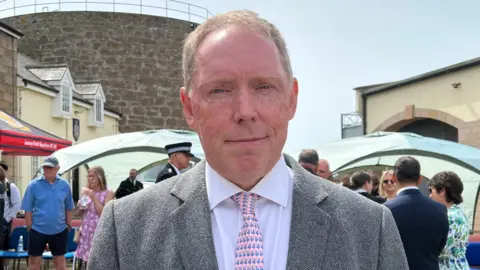 Paul Smith in a grey suit and pink tie standing in a courtyard, with a crowd, chairs, and event tents set up behind them.