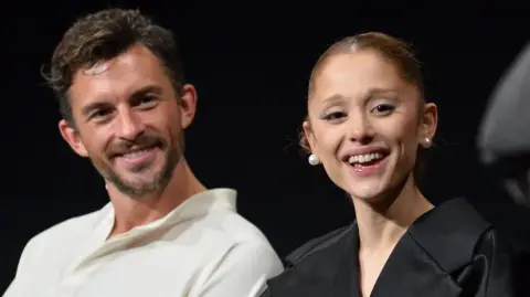 Getty Images Jonathan Bailey and Ariana Grande seated together and smiling during a Wicked promotional event in November 2025