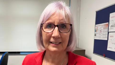 A woman with silvery pink hair and glasses smiles into the camera. 